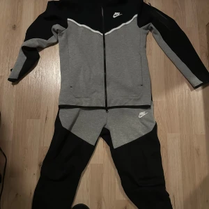 Nike grå och svart zip hoodie - Säljer en snygg grå och svart hoodie från Nike med hel dragkedja och vit logga på bröstet. Tröjan har lång ärm, sportig design och är tillverkad i mjukt bomullsmaterial. Perfekt för träning eller chill.
