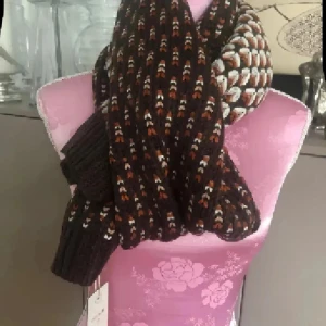 Brun stickad halsduk med mönster - Snygg och bred halsduk i stickad kvalitet med brunt, vitt och beige mönster. Mönstret är randigt och geometriskt, vilket ger en cool retrovibe. Perfekt att svepa runt halsen för extra värme och stil under kyliga dagar.ny med prislappen från danska Cream 