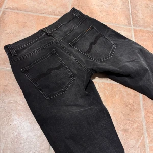 Svarta jeans från Nudie Jeans, 32/30 - Snygga svarta jeans från Nudie Jeans i klassisk femficksmodell. Jeansen har en rak passform med diskret slitning och Nudie Jeans signaturbroderi på bakfickorna. Materialet är mjukt denim och midjan är normal. Perfekt för dig som gillar stilrena och tidlösa jeans.                           Modell: Grim tim. 
