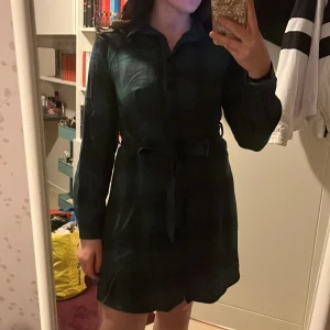 Mörkgrön kort skjortklänning med bälte - Snygg mörkgrön kort skjortklänning med långa ärmar och krage. Klänningen har ett matchande bälte i midjan som knyts vid midjan. 