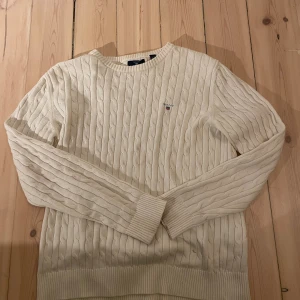 Beige kabelstickad tröja från GANT - Snygg vit/beige kabelstickad tröja från GANT med rund hals och broderad logga på bröstet. Tröjan har långa ärmar och breda ribbade muddar vid ärmslut och nederkant. Perfekt för dig som gillar klassisk och clean stil.Passar om man är 170 ungefär