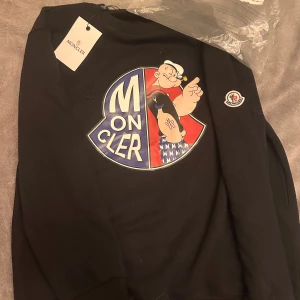 Svart Moncler sweatshirt med Popeye - Svart sweatshirt från Moncler med stor färgglad Popeye-tryck på bröstet och Moncler-logga på ärmen. Rund hals och ribbade muddar. Perfekt för dig som gillar streetstyle och vill sticka ut med en unik design. Strl Xs/S. R3pl1c4