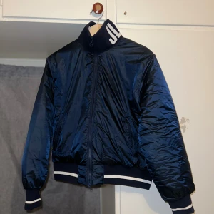 J.Lindeberg Bomberjacka - Mörkblå bomberjacka från J.Lindeberg med glansig finish och vita JL-loggor på kragen. Jackan har ribbade muddar med vita ränder, dragkedja framtill och quiltad insida. Perfekt för dig som vill ha en sportig och stilren look.