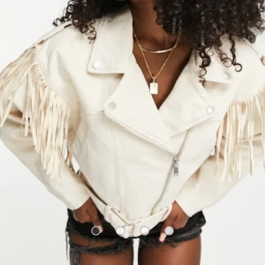 Beige jacka med fransar - Säljer en beige jacka med coola fransar längs axlarna och ärmarna. Jackan har sned dragkedja, bred krage och bälte i midjan. Oversized för en avslappnad vibe. Kan inte se någon storlek, men passar mig som brukar ha XS/S