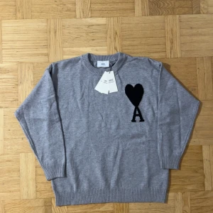 Grå Ami Paris Tröja  - Helt ny och aldrig ens använd. Säljs eftersom den är väldigt liten för mig. Snygg grå tröja från Ami Paris i ekologisk bomull. På bröstet finns en svart hjärtdetalj med bokstaven A. Tröjan har rund hals, ribbade muddar och lång ärm. Perfekt för dig som gillar stilrena plagg med en twist. Perfekt nu när vintern kommer. För fler bilder skriv. 