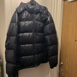 Mörkblå pufferjacka Polo Ralph Lauren - Mörkblå pufferjacka från Polo Ralph Lauren med huva och dragkedja framtill. Jackan har en diskret röd logga på bröstet, flera fickor med dragkedja och en glansig finish. Perfekt för kalla dagar och riktigt snygg i vintermörkret. Storlek L men lite större i sin size så snarare att den funkar som L/XL.