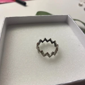 Zigzag silverring med stenar - Cool ring i silverfärg med ett unikt zigzag-mönster runt hela ringen. Små glittrande stenar sitter längs kanterna och ger extra bling. Perfekt för dig som gillar att sticka ut med dina accessoarer. Passar både till enkel och edgy stil.
