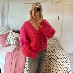 Rosa kofta från & Other Stories - Superfin rosa kofta från & Other Stories med grovstickat kabelmönster, v-ringning och stora matchande knappar. Modellen är något croppad med puffiga långärmar och breda muddar.