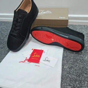 Svarta sneakers från Christian Louboutin - Snygga svarta sneakers från Christian Louboutin med klassisk röd sula. Skorna har rund tå, platt sula och är tillverkade i mocka med svarta snören. Insidan är beige och har märkets logga i guld. Perfekt för dig som gillar exklusiv och stilren design.