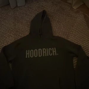 Svart hoodie från Hoodrich - Svart hoodie från Hoodrich med stor vit logga tryckt över bröstet. Tröjan har huva, lång ärm och en klassisk magficka framtill. Materialet är mjukt och känns som bomull, perfekt för chill dagar. Snygg och enkel streetwear-stil.
