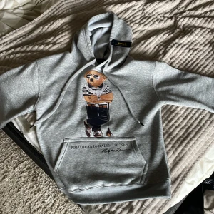 Grå hoodie Polo Bear från Ralph Lauren - Säljer en grå hoodie från Polo Ralph Lauren i storlek S med Polo Bear-tryck på framsidan. Tröjan har huva med snörning, magficka och ribbade muddar. Materialet är mjukt och känns som bomull och polyester. Tyvärr så är den för liten för mig, använd 1-2 ggr för att enbart testa den. Orginal pris är 3500+ så detta pris är verkligen generöst. Vid en snabb affär kan priset gå ner lite. Tveka inte att höra av för frågor och mer