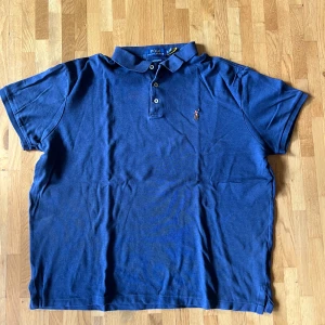 Blå pikétröja från Polo Ralph Lauren XL - Säljer en klassisk blå pikétröja från Polo Ralph Lauren i modellen Custom Slim Fit. Tröjan har korta ärmar, krage och knappar framtill samt den ikoniska broderade polospelaren på bröstet. Perfekt för en stilren och avslappnad look.