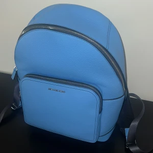 Blå ryggsäck i skinn från Michael Kors - Snygg blå ryggsäck från Michael Kors i äkta skinn. Väskan har en stilren design med silverfärgade dragkedjor, justerbara axelband och en praktisk ytterficka framtill. Perfekt för dig som vill ha en trendig och funktionell väska till vardagen.
