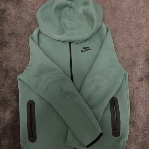 Nike grön hoodie Tech Fleece XL - Nike Tech Fleece hoodie i en snygg grön färg med svarta detaljer och dragkedjor på fickorna. Tröjan har huva, lång ärm och klassisk Nike-logga på bröstet. Perfekt för chill eller träning, riktigt skön och stilren modell . Köpt för 877 kr. ( st : XL  158 - 170 ( barn storlek )