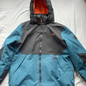 Blå och svart vindjacka från North Bend - Vindjacka från North Bend i blått och svart med orange detaljer och huva. Jackan har hel dragkedja, vattentäta sömmar och orangefärgat foder. Perfekt för blåsiga och regniga dagar, med sportig look och praktiska funktioner.