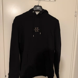 Svart hoodie från Les Deux - Svart hoodie från Les Deux med broderad logga på bröstet och klassisk känguruficka. Tröjan har huva med snörning och långa ärmar. Perfekt för en chill och stilren look.