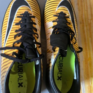 Nike Mercurial X gula fotbollsskor - Säljer ett par Nike Mercurial X futsal skor i en riktigt cool gul och svart design med vita detaljer. Skorna har snörning, platt sula och är tillverkade i syntetmaterial. Perfekta för dig som vill sticka ut på planen med stilrena och sportiga skor.
