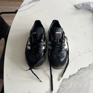 Adidas Samba sneakers svart. - Säljer ett par klassiska Adidas Samba sneakers i svart med vita ränder och brun gummisula.  Dessa skor köptes för en månad sedan och kostade cirka 1400. Skorna har används i en vecka inomhus men passade inte riktigt då tyckte dom satt lite tajt så köpte en storlek större och dom passade perfect. Priset kan diskuteras om skorna och det är bara att höra av sig.