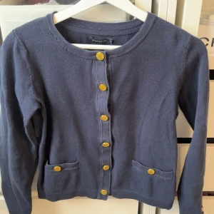 Mörkblå kofta från Stockh LM - Snygg mörkblå kofta från Stockh LM med guldfärgade knappar framtill och på fickorna. Koftan har rund halsringning, två små fickor och är långärmad. Materialet känns mjukt och är stickat, perfekt för lager på lager under kyliga dagar.