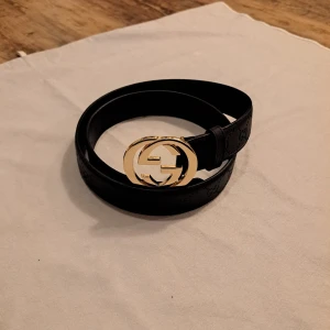 Svart bälte med Guld spänne - Säljer ett svart bälte med präglade GG-loggor längs hela bandet. Skärpet har ett stort, rundat spänne i guld med dubbla G:n. Perfekt accessoar för att lyfta vilken outfit som helst.