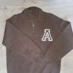 Brun stickad polotröja med A-märke - Snygg brun stickad polotröja med halv dragkedja och stor vitbrun A-applikation på bröstet. Tröjan har ribbade muddar och krage, perfekt för dig som gillar college-vibe och vill ha något varmt och chill. Passar dig som vill sticka ut med en cool detalj.