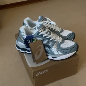 Asics Gel-Kayano 14 sneakers vit/blåa - Fräscha Asics Gel-Kayano 14 sneakers i vit och blå med silvriga detaljer. Skorna har meshpaneler för bra ventilation, snörning och en chunky sula med gel-dämpning. Perfekt för dig som gillar sportig stil och komfort. Ortholite-innersula för extra stöd.