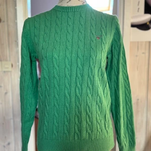 Grön kabelstickad tröja från GANT - Snygg grön kabelstickad tröja från GANT med rund halsringning och diskret logga på bröstet. Tröjan har långa ärmar och ribbade muddar vid ärmslut och nederkant. 