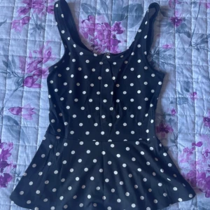 Svart prickig baddräkt med dragkedja - Supersöt svart baddräkt med vita prickar och en snygg dragkedja i ryggen. Modellen har breda axelband och en liten volangkant nedtill som ger en retrovibe. Perfekt för stranden eller poolen i sommar!