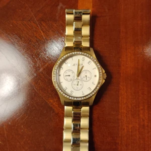 Jet Set guld klocka med kristaller - Snygg Jet Set klocka i guld med länkarmband i rostfritt stål. Urtavlan är ljus med tre mindre visartavlor och dekorerad med kristaller runt boetten. Jet Set-loggan syns både på urtavlan och spännet. Perfekt accessoar för dig som gillar bling och stil.