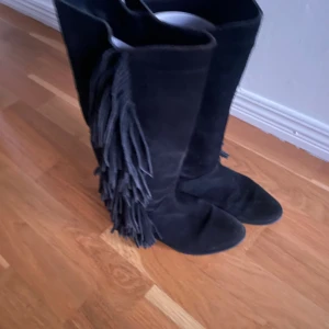 Svarta mockastövlar med fransar - Säljer ett par svarta höga stövlar i mocka med snygga fransdetaljer längs utsidan. Skorna har spetsig tå, vilket ger en cool och avslappnad vibe. Perfekta för dig som vill sticka ut lite extra.