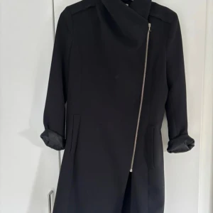 Svart kappa med sned dragkedja - Stilren svart kappa med sned dragkedja framtill och dragkedjor vid ärmsluten. Jackan har en rak och något figursydd passform, klassisk hög krage och diskreta fickor. Perfekt för dig som gillar minimalistisk och modern stil.