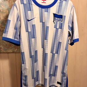 Hertha BSC 2021/22 Nike home shirt, size L - Säljer en officiell Hertha BSC matchtröja från Nike i storlek L. Tröjan är vit med blå randigt mönster och har klubbmärket på bröstet samt sponsortryck på ärmen. Tillverkad i lätt Dri-Fit material som andas, perfekt för träning eller match.