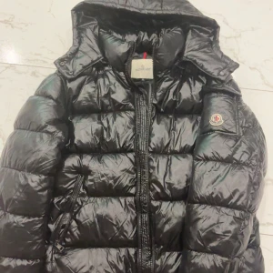 Svart Moncler pufferjacka med huva - Säljer en svart pufferjacka från Moncler med ikonisk logga på ärmen och huva. Jackan har quiltad design, dragkedja framtill och sidofickor med dragkedja. Insidan har en cool serietidningsetikett. Perfekt för dig som vill ha en snygg och varm vinterjacka.Den kan va lite svårt att stänga pågrund den är ny och inte använd 