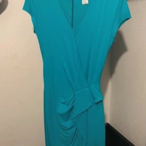 Turkos omlottklänning med knyt - Snygg turkos klänning med omlottkänsla och knyt i midjan. Klänningen har v-ringning och korta ärmar, vilket ger en fräsch och modern look. Perfekt för dig som vill sticka ut med färg och stil.