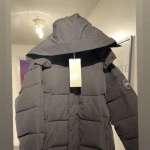 Svart pufferjacka från Canada Goose - Säljer en svart pufferjacka från Canada Goose med stor huva och patch på ärmen. Jackan har quiltad design, dragkedja och knappar framtill samt två sidofickor. Perfekt för kalla dagar och har en clean, modern look.