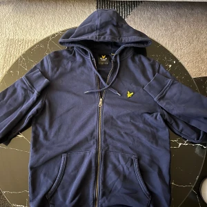 Marinblå hoodie från Lyle & Scott - Snygg marinblå hoodie från Lyle & Scott med gul broderad logga på bröstet. Tröjan har huva med snörning, dragkedja framtill och två fickor. Tillverkad i mjuk bomull, perfekt för chill dagar. Klassisk och stilren design som funkar året runt.