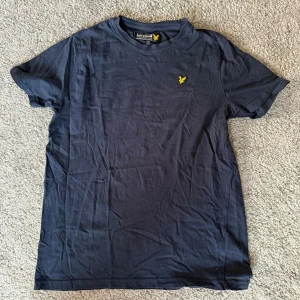 Mörkblå t-shirt från Lyle & Scott - Snygg mörkblå t-shirt från Lyle & Scott med klassisk gul logga broderad på bröstet. T-shirten har rund hals och korta ärmar, tillverkad i mjuk bomull som är skön mot huden. Perfekt för dig som gillar stilrena och enkla plagg.