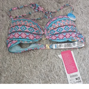 Färgglad bikiniöverdel med mönster - Säljer en bikiniöverdel med färgstarkt geometriskt mönster i rosa, blått och svart. Modellen har snörning i nacken och ett snyggt guldfärgat spänne bak. Perfekt för stranden och sommarens soliga dagar!