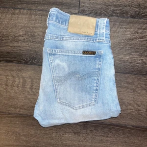 Nudie jeans  - Säljer ett par ljusblå skinny jeans från Nudie Jeans i storlek 26/34 sitter mer som L30.. Jeansen har klassisk femficksdesign, snygga slitningar och Nudie-logga på bakfickan. Modell Tight Long John. Midja 33cm, längd 91cm. JAG SKICKAR EJ FLER BILDER NÄR JAG BÄR JEANSEN! för storleksguide hänvisar jag till måtten och för bättre inblick i passformen rekommenderar jag att googla på modellnamnet. Jeansen kan vara uppsydda utan att jag noterat det så kolla måtten noggrant :) s3