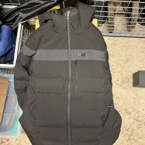 Svart pufferjacka Helly Hansen M - Svart pufferjacka från Helly Hansen med huva och hel dragkedja. Jackan har en mörkgrå horisontell rand över bröstet och ryggen samt HH-logga på bröstet. Regular fit, perfekt för kalla dagar. Snygg och stilren design med praktiska fickor.