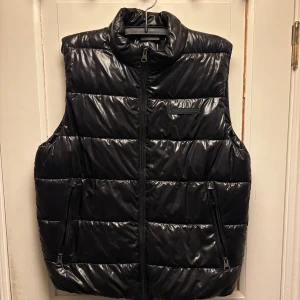 Svart dunväst med dragkedja - Säljer en svart dunväst med glansig yta och hög krage. Västen har två sidofickor med dragkedja och en bröstficka med dragkedja. Perfekt att slänga över en hoodie när det är kyligt ute. Stängs med en kraftig dragkedja framtill. 