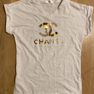 Vit t-shirt med guldtryck från Tina - Vit t-shirt med v-ringning och korta ärmar från Tina. Framsidan har ett stort Chanel-inspirerat tryck i guld och små glittriga detaljer. Materialet känns som mjuk bomull och passformen är normal. Perfekt för dig som gillar statement-plagg och lyxig vibe.
