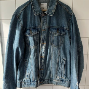 Blå jeansjacka från Bershka - Klassisk blå jeansjacka från Bershka med snygga slitningar och kontrastsömmar. Jackan har två bröstfickor med knappar, två sidofickor och metallknappar framtill. Perfekt oversized fit och tidlös denim-look.