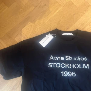 Svart Acne Studios t-shirt S Stockholm - Svart t-shirt från Acne Studios med vit text 'Acne Studios STOCKHOLM 1996' på bröstet. Klassisk rund hals och korta ärmar. Materialet är mjuk bomull och passformen är normal. Perfekt för dig som gillar stilrena och ikoniska plagg med streetkänsla.