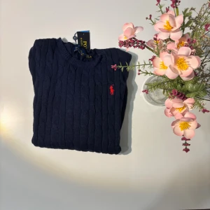 Marinblå stickad tröja Polo Ralph Lauren - Snygg marinblå stickad tröja från Polo Ralph Lauren med klassisk kabelstickning och röd broderad logga på bröstet. Rund halsringning och långa ärmar. Perfekt för dig som gillar stilrena och tidlösa plagg med premiumkänsla.