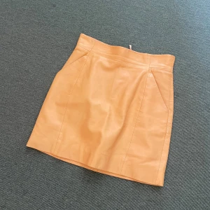 Beige skinnkjol från H&M, strl 34 - Snygg beige kort skinnkjol från H&M i storlek 34. Kjolen har en stilren design med hög midja, diskreta sömmar och dragkedja bak. Perfekt för dig som vill ha en trendig och clean look.