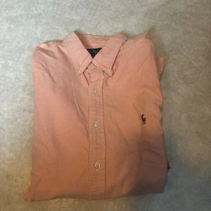 Rosa skjorta från Polo Ralph Lauren - Snygg rosa skjorta från Polo Ralph Lauren i slim fit-modell. Skjortan har klassisk krage, knappar hela vägen och det ikoniska broderade Polo-logot på bröstet. Tillverkad i mjuk bomull och har långa ärmar med manschettknappar. Perfekt för dig som gillar stilrena och fräscha plagg.