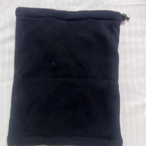 Svart fleecehalsduk med grå pälsfoder - Mysig svart halsduk i fleece med fluffigt grått pälsfoder på insidan. Tubmodell med dragsko upptill som gör att den sitter extra bra runt halsen. Perfekt för kalla dagar när du vill hålla dig varm och bekväm.