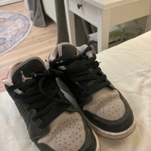 Nike Air Jordan 1 Low svart/rosa - Nike Air Jordan 1 Low sneakers i svart och rosa med klassisk Jumpman-logga på tungan och Jordan Wings broderat på hälen. Skorna har snörning, perforerad tå och platt sula. Materialet är skinn och syntet, med rund tå och lågt skaft.