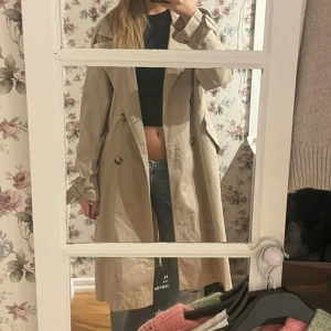 Beige trenchcoat från Bershka - Stilren beige trenchcoat från Bershka med klassisk dubbelknäppning, bred krage och bälte i midjan. Jackan har långa ärmar med spännen vid handlederna och stora fickor framtill. Köptes på Sellpy i nyskick med lapp, men endast använd en gång av mig!